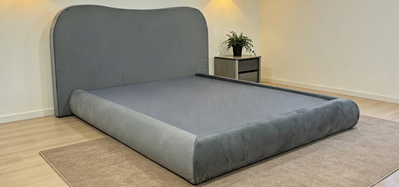Close-up of the gray upholstered bed frame's base and headboard set against a neutral wall in a real-life room setting with a wooden floor.
